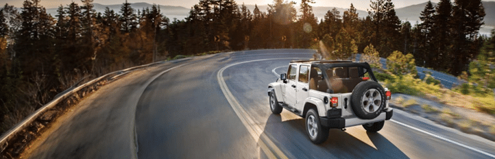A white SUV driving on a scenic winding road through a forest, with a view of distant mountains at sunset.