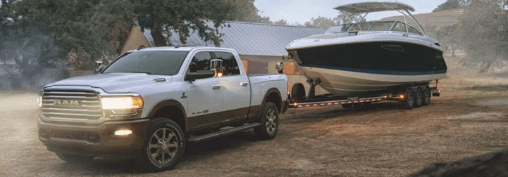 A pickup truck towing a large boat on a trailer in a rural setting with trees and a house nearby.
