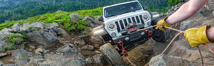 Off-road vehicle being winched uphill on rocky terrain by a person in gloves, surrounded by greenery.