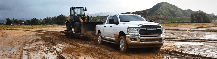 A white pickup truck towing a construction vehicle on a muddy, rural road with hills in the background.