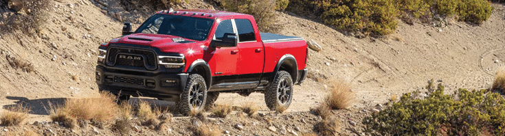 Red pickup truck driving on a rugged, sandy off-road terrain surrounded by sparse vegetation.