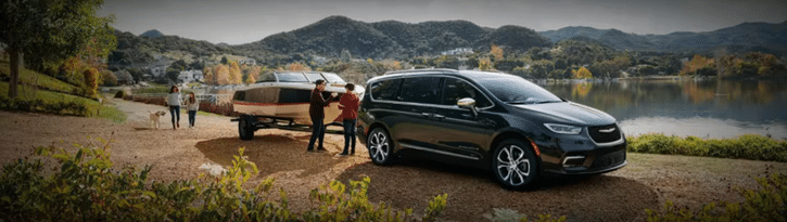 Minivan towing a boat on a scenic lakeside dirt road with hills in the background.