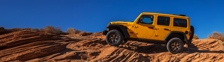 Yellow Jeep Rubicon climbing a red rocky slope under a clear blue sky.