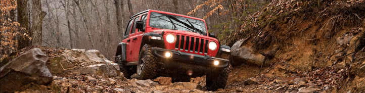 Red off-road vehicle navigating a rugged, rocky trail in a wooded area.