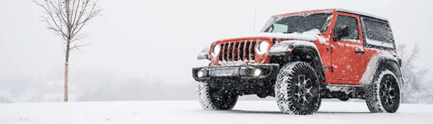 Red off-road SUV driving on a snowy terrain, headlights on, with a leafless tree nearby.
