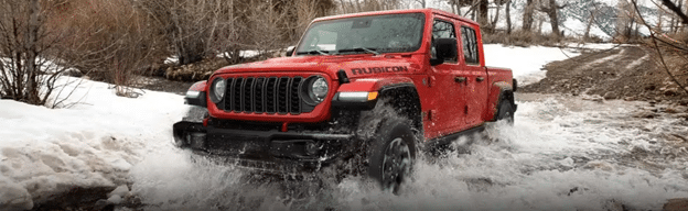 Red Rubicon truck driving through a snowy, watery terrain with surrounding bare trees.