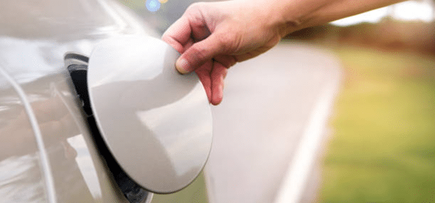 Hand opening car fuel cap, with blurred road and greenery in the background.