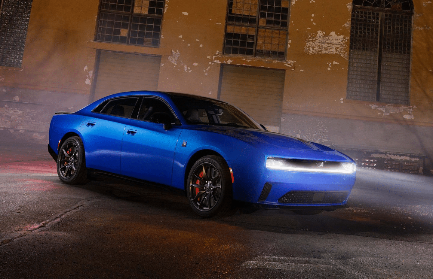 Blue sports car with illuminated headlights parked in front of an industrial building at night.