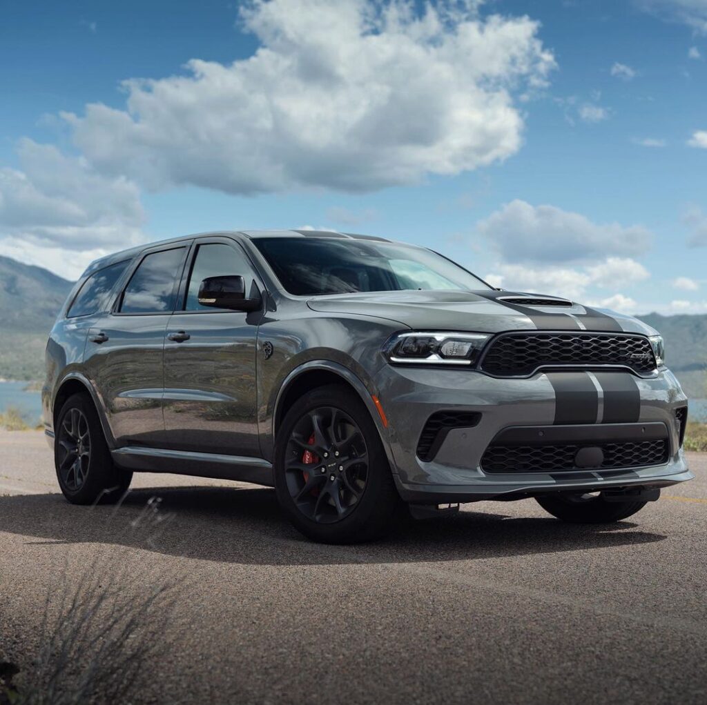 Silver SUV with black racing stripes parked on a road, with mountains and blue sky backdrop.
