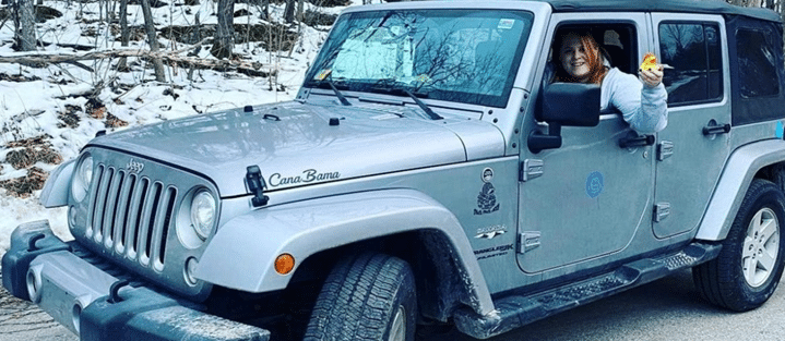 Woman in a silver Jeep Wrangler on a snowy road, holding a small yellow rubber duck out the window.
