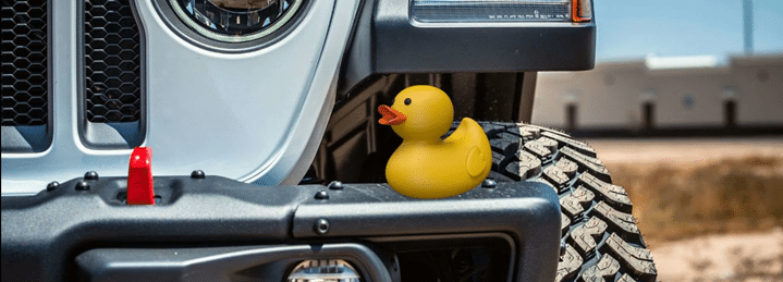 Yellow rubber duck on a Jeep bumper near tire, with off-road terrain in the background.