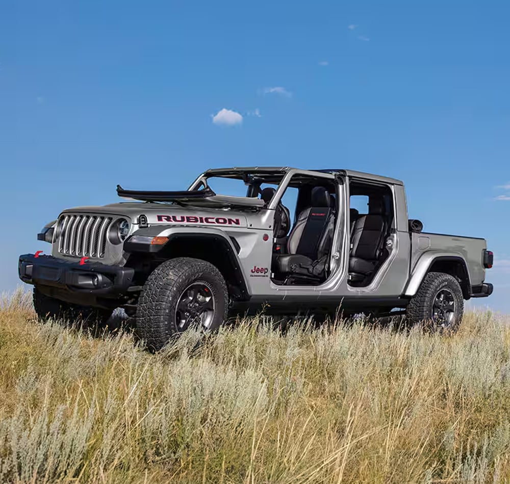Silver Jeep Rubicon with open doors on a grassy field under a blue sky.