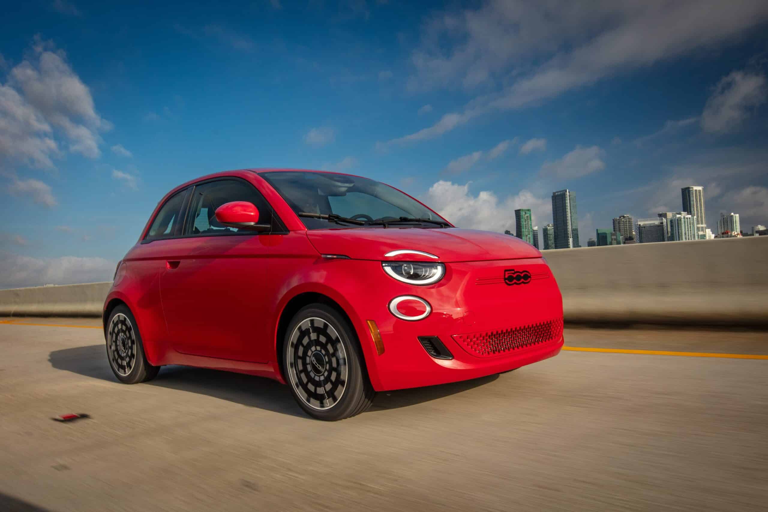 Red compact car driving on a highway with a city skyline in the background under a blue sky.