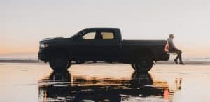 Silhouetted pickup truck at sunset on a wet beach with a person seated on the tailgate.
