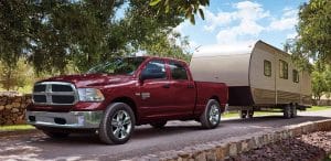 A red pickup truck towing a beige camper trailer on a tree-lined suburban road.